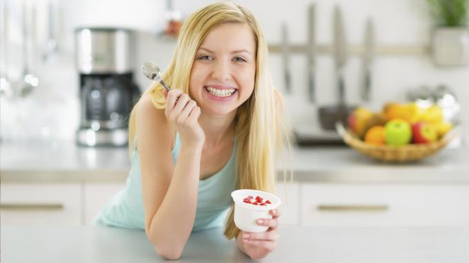 Laimīga jauna sieviete virtuvē ēd jogurtu