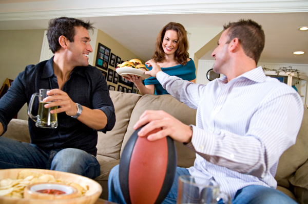 Жена послужује хамбургере мужу и пријатељу који гледају фудбал